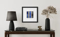 A framed modern abstract art print inspired by stained-glass church windows featuring a blue, gray, and black plaid pattern, displayed on a white wall above a dark wood console table styled with a black lamp, books, and a vase of dried flowers.