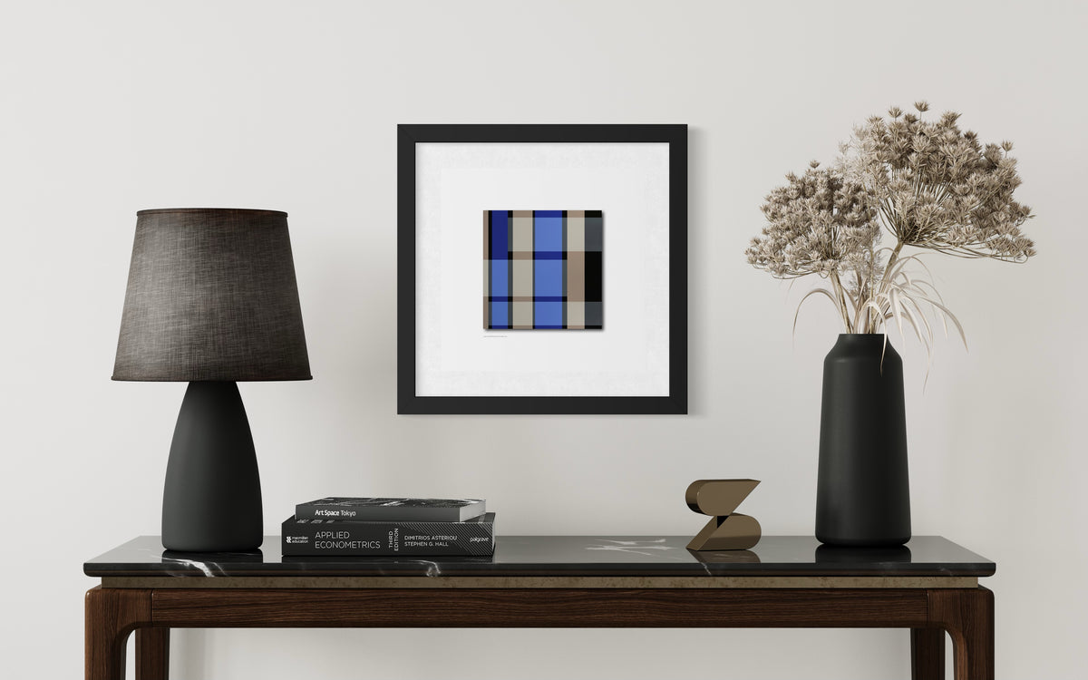 A framed modern abstract art print inspired by stained-glass church windows featuring a blue, gray, and black plaid pattern, displayed on a white wall above a dark wood console table styled with a black lamp, books, and a vase of dried flowers.