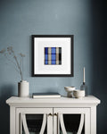 A framed modern abstract art print inspired by church windows featuring a blue, black, and gray plaid pattern, displayed on a textured blue-gray wall above a white sideboard with decorative ceramic vases and bowls.