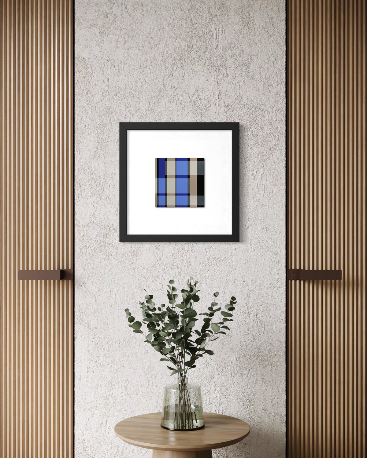 A framed modern abstract art print inspired by church windows featuring a blue and gray grid pattern, displayed on a textured light gray wall above a small wooden table with a glass vase of green leaves.