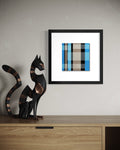 A framed abstract modern art print featuring a teal blue, gray, and beige plaid pattern, displayed on a light gray wall above a wooden console table next to a black cat sculpture.