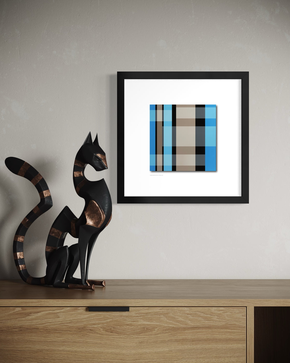 A framed abstract modern art print featuring a teal blue, gray, and beige plaid pattern, displayed on a light gray wall above a wooden console table next to a black cat sculpture.