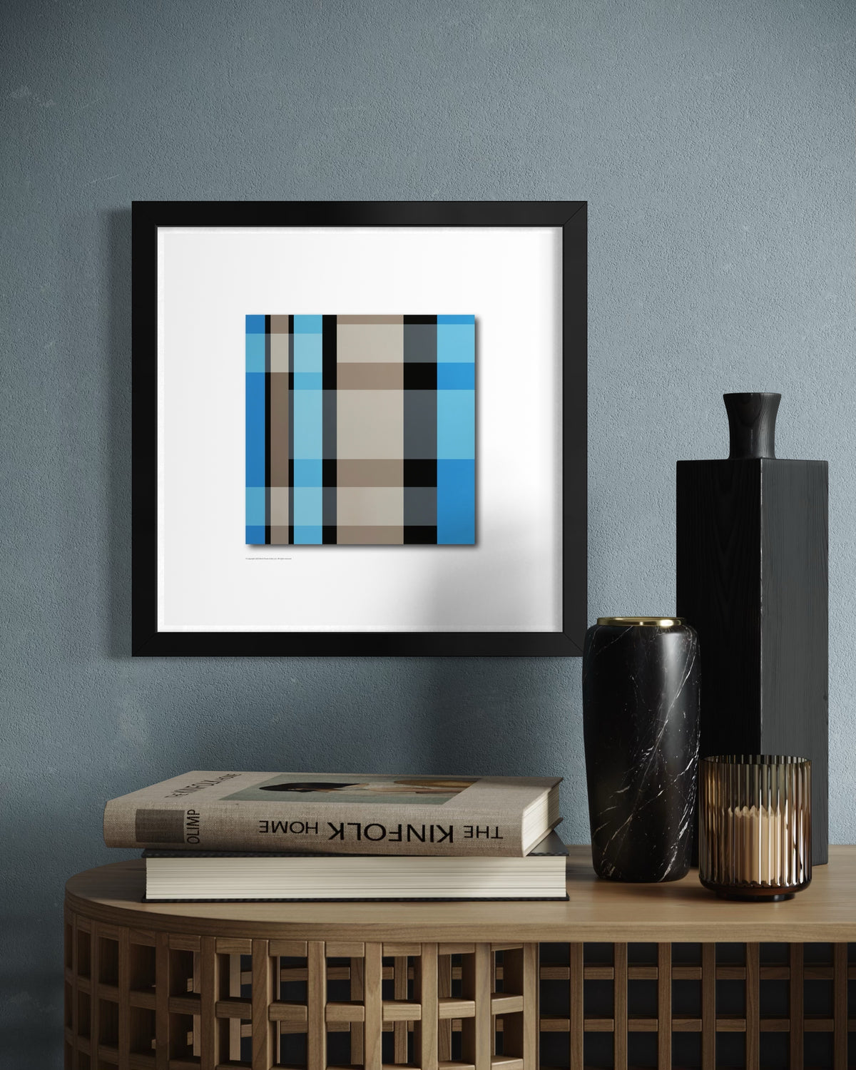 Framed contemporary, modern abstract wall art based on stained-glass windows with vertical stripes in teal blue, black, beige, and white, displayed in a black frame on a textured blue-gray wall above a wooden side table with books and decorative vases.