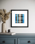 A framed abstract stained-glass-inspired art print with teal blue, beige, and black stripes, displayed on a white wall above a blue-gray console table with a vase of pampas grass and a book.