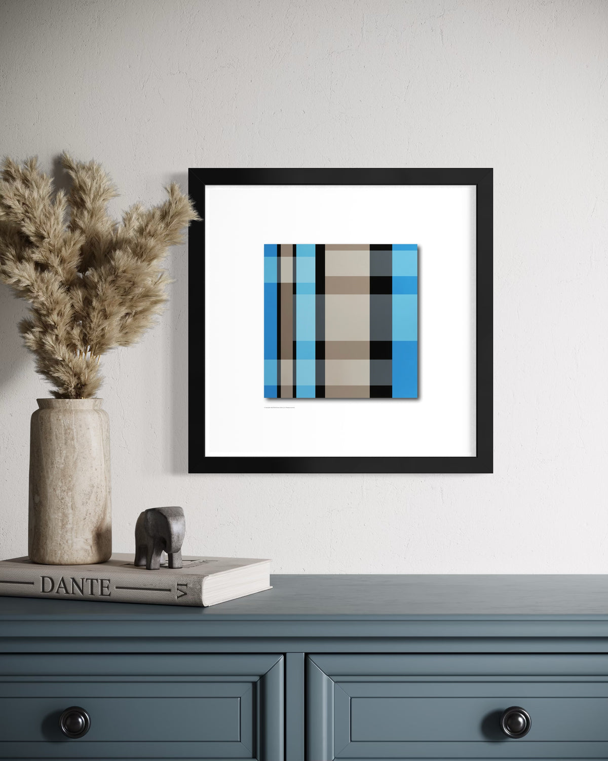A framed abstract stained-glass-inspired art print with teal blue, beige, and black stripes, displayed on a white wall above a blue-gray console table with a vase of pampas grass and a book.
