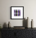 A framed abstract contemporary art print with a spiritual story featuring a purple, black, and beige plaid pattern, displayed on a light gray wall above a dark textured cabinet with decorative vases and books.