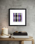 A framed abstract modern art print with a spiritual story featuring a purple, black, gray, and white plaid pattern, displayed on a textured gray wall above a wooden table with books and a candle.