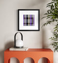 A framed art print featuring a purple, gray, and black plaid pattern, displayed on a white wall above a modern orange side table with a white lamp and green plant leaves.