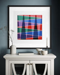 A framed abstract geometric art print inspired by mountains and shadows in the desert with intersecting red, green, blue, purple, and black lines, displayed in a black frame on a dark blue-gray wall above a white console table with decorative objects.