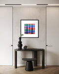A framed colorful geometric art print premised on mountains and shadows with colorful intersecting lines hangs on a textured light gray wall above a dark console table and black stool in a modern interior setting.