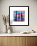 Using desert life and mountains as a theme, this framed museum-quality abstract art print hangs above a console table, featuring intersecting diagonal bands in red, blue, green, purple, and pink on a white mat. The black frame is shown in a styled interior setting with a vase, small ceramic vessel, open book, and bowl on a marble surface.