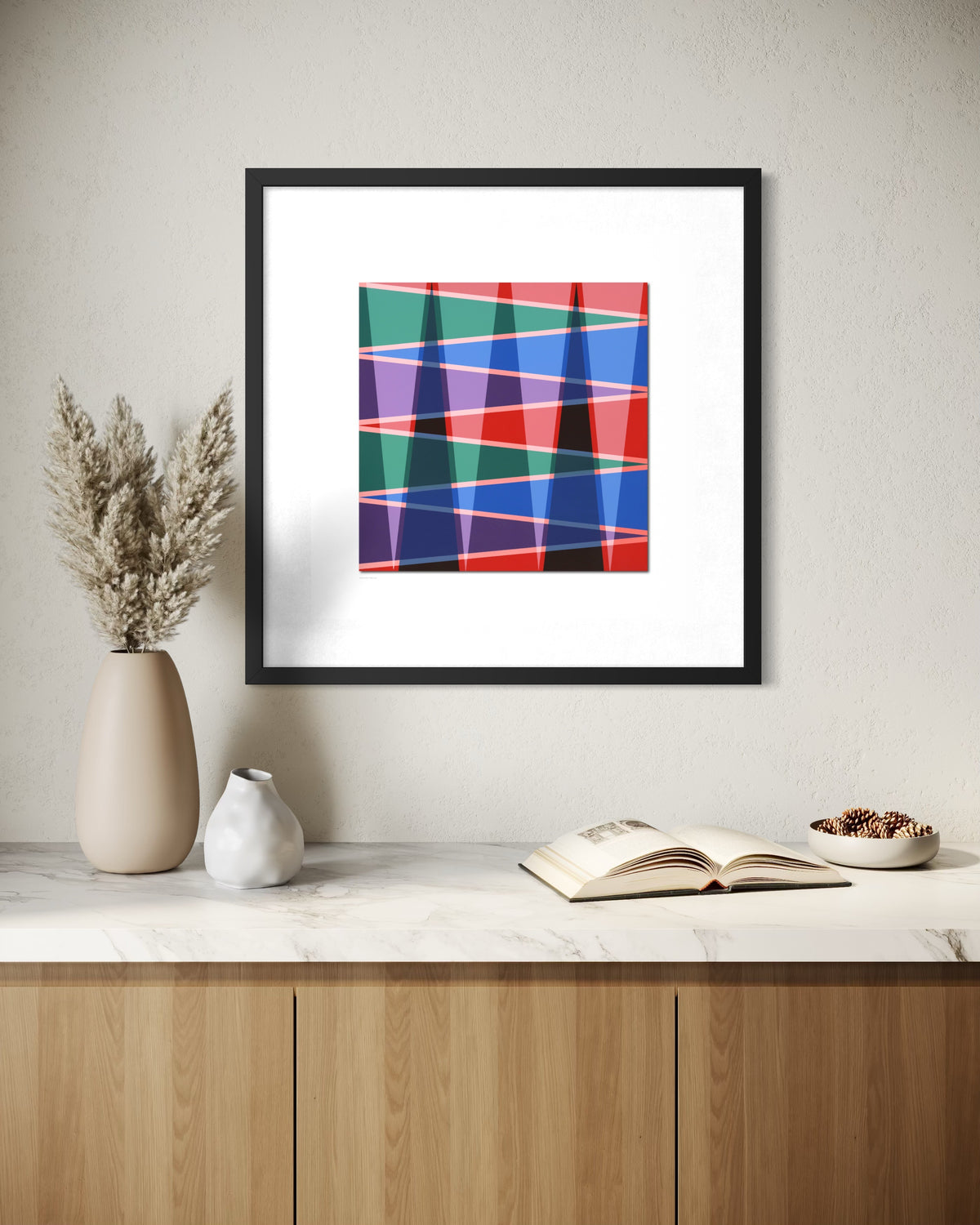Using desert life and mountains as a theme, this framed museum-quality abstract art print hangs above a console table, featuring intersecting diagonal bands in red, blue, green, purple, and pink on a white mat. The black frame is shown in a styled interior setting with a vase, small ceramic vessel, open book, and bowl on a marble surface.
