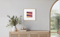 A framed abstract desert-scape art print with diagonal stripes in purple, orange, and yellow, displayed on a light gray wall above a wooden console table with a vase of greenery.