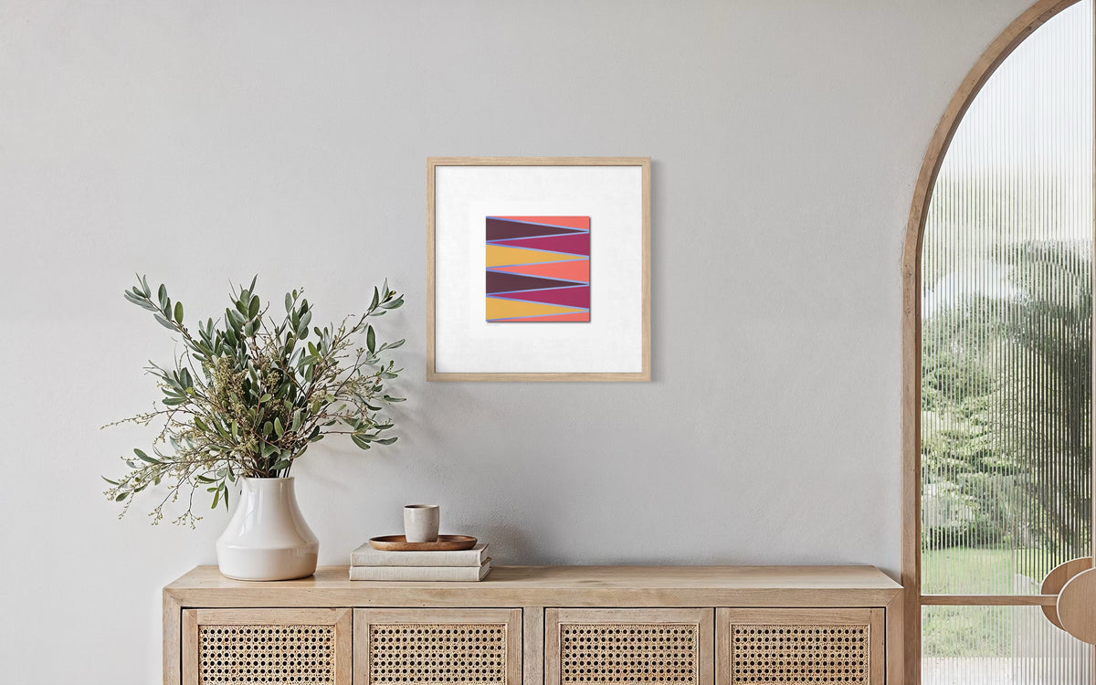 A framed abstract desert-scape art print with diagonal stripes in purple, orange, and yellow, displayed on a light gray wall above a wooden console table with a vase of greenery.