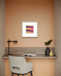 A framed abstract geometric desert-scape art print with pink, purple, orange, and yellow stripes, displayed on a peach-colored wall above a modern gray desk setup with a chair, lamp, and vases.