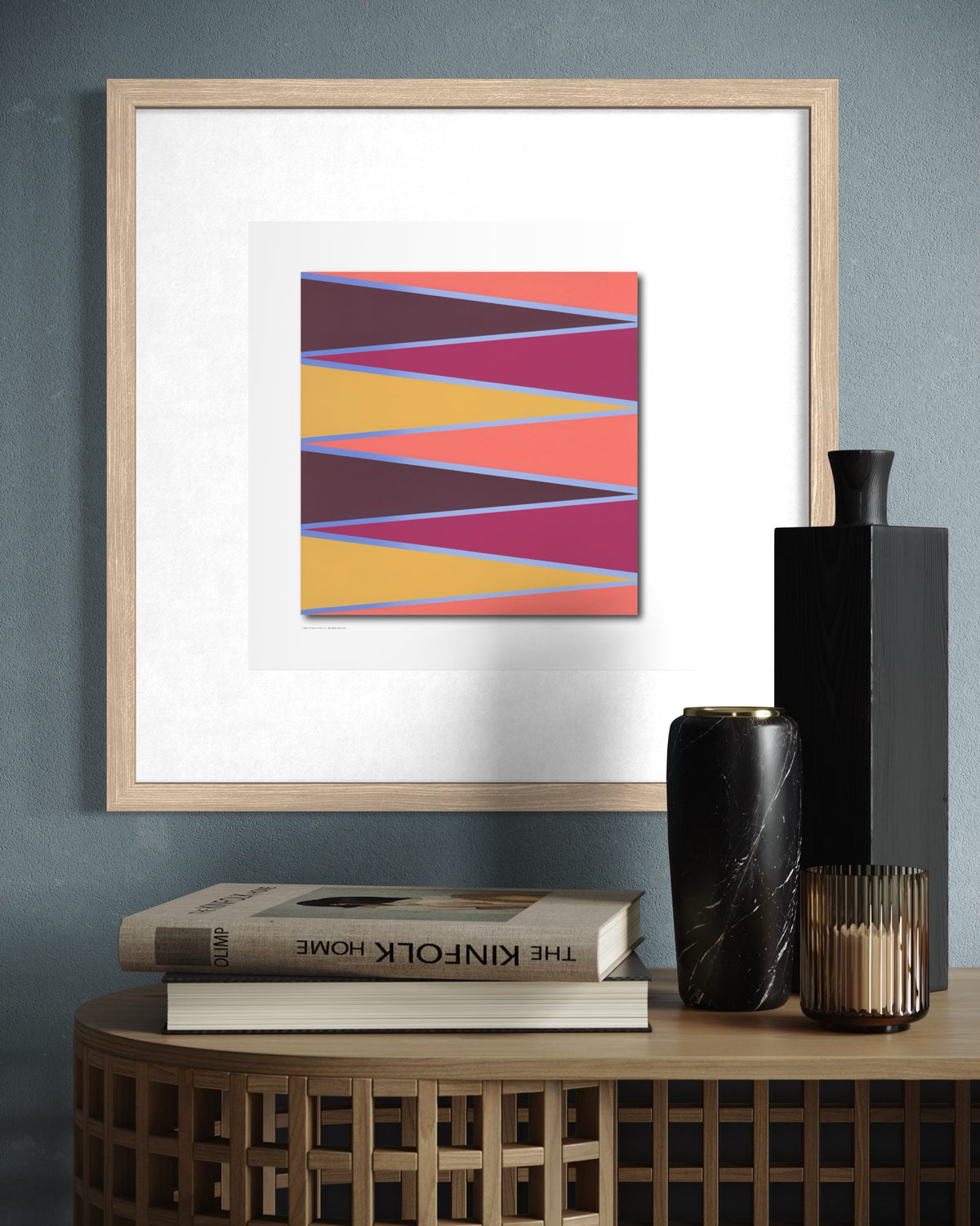 Framed abstract desert-scape geometric wall art with colorful diagonal stripes in pink, orange, purple, and yellow, displayed in a light wood frame on a gray wall above a wooden console table with books and vases.