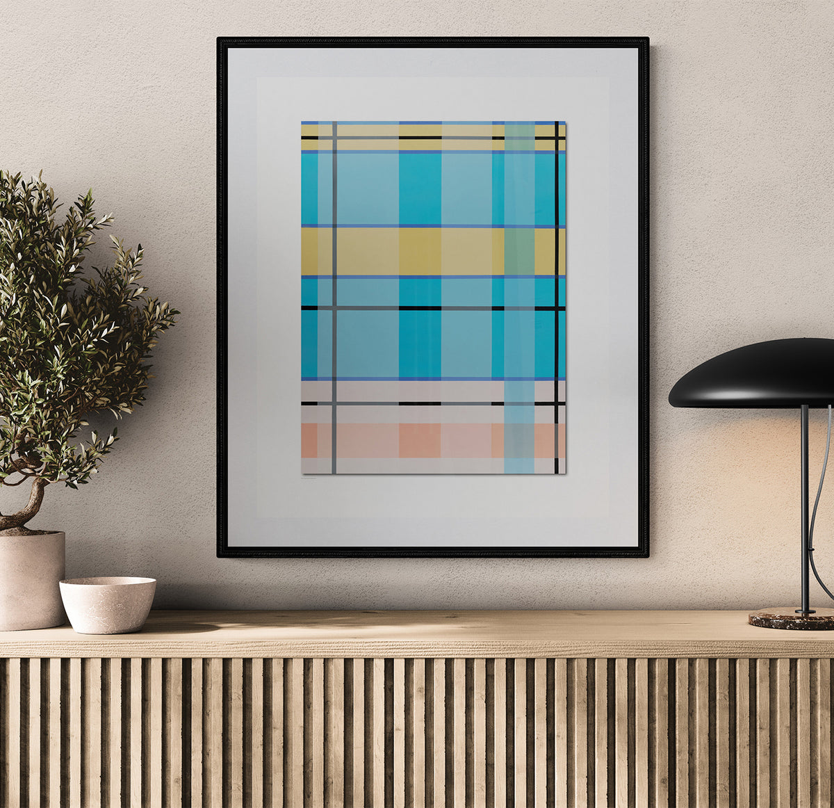 A framed abstract geometric art print with blue, yellow, and pink plaid-like blocks hangs on a beige wall above a wooden console table, styled with a potted plant and a black table lamp.