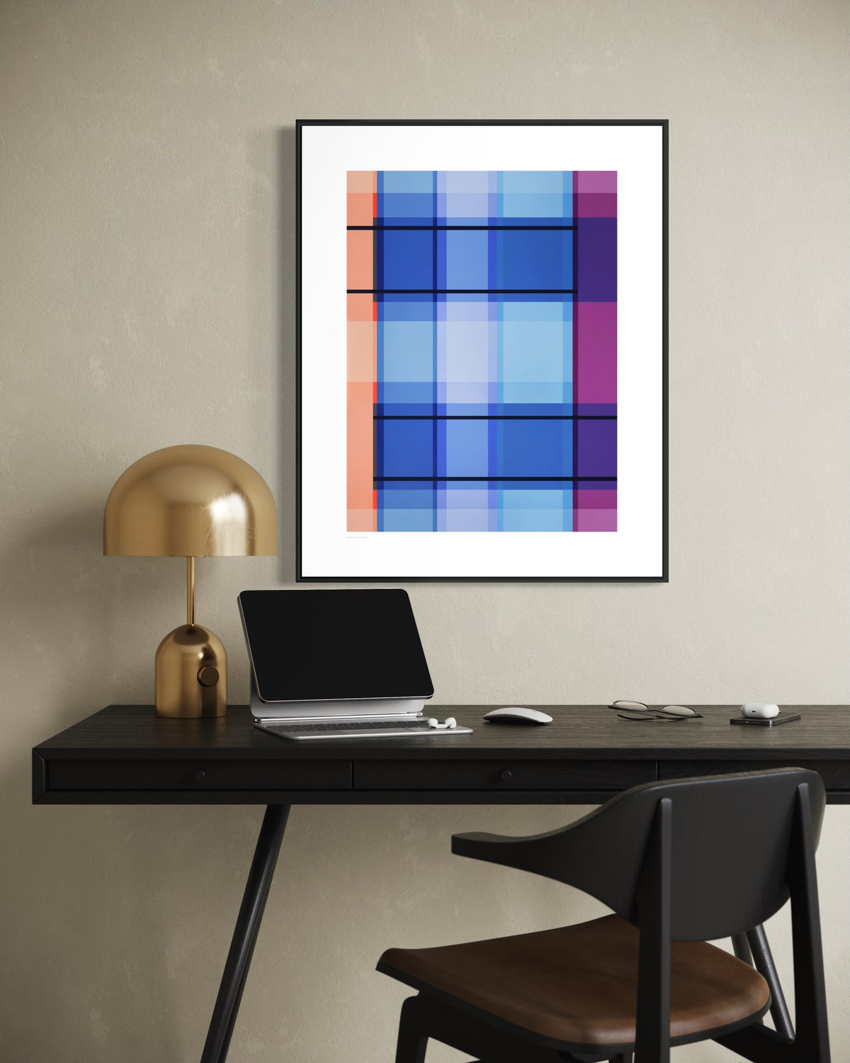A framed contemporary geometric art print with blue, purple, and orange blocks hangs on a textured beige wall above a dark desk with a laptop, mouse, and a gold desk lamp. A black chair with a brown leather seat is in the foreground.