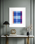A framed contemporary abstract art print featuring a geometric plaid pattern in shades of blue, purple, and orange, displayed on a gray paneled wall above a console table with a lamp, books, and a vase.