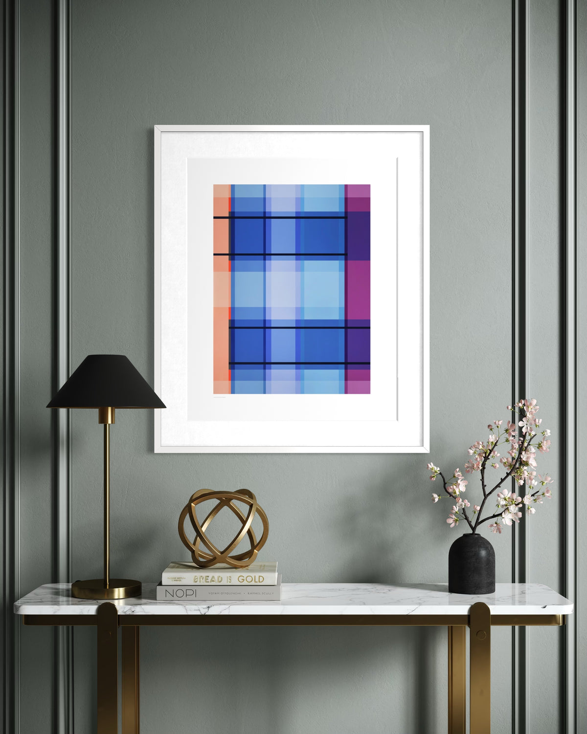 A framed contemporary abstract art print featuring a geometric plaid pattern in shades of blue, purple, and orange, displayed on a gray paneled wall above a console table with a lamp, books, and a vase.