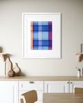 A framed contemporary abstract geometric art print with a plaid-like grid pattern in shades of blue, purple, and orange, displayed in a white frame on a beige wall above a kitchen counter.