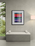 A framed abstract plaid art print with vibrant red, blue, green, and purple stripes, displayed on a textured gray wall above a modern gray bench in a minimalist room setting.