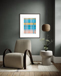 A framed abstract geometric art print with blue, yellow, and pink plaid-like blocks hangs on a dark gray wall above a beige armchair and a small side table with a vase of flowers.