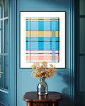 A framed abstract geometric art print with blue, yellow, and pink plaid-like blocks hangs on a teal wall above a dark wood console table with a silver vase of dried flowers.