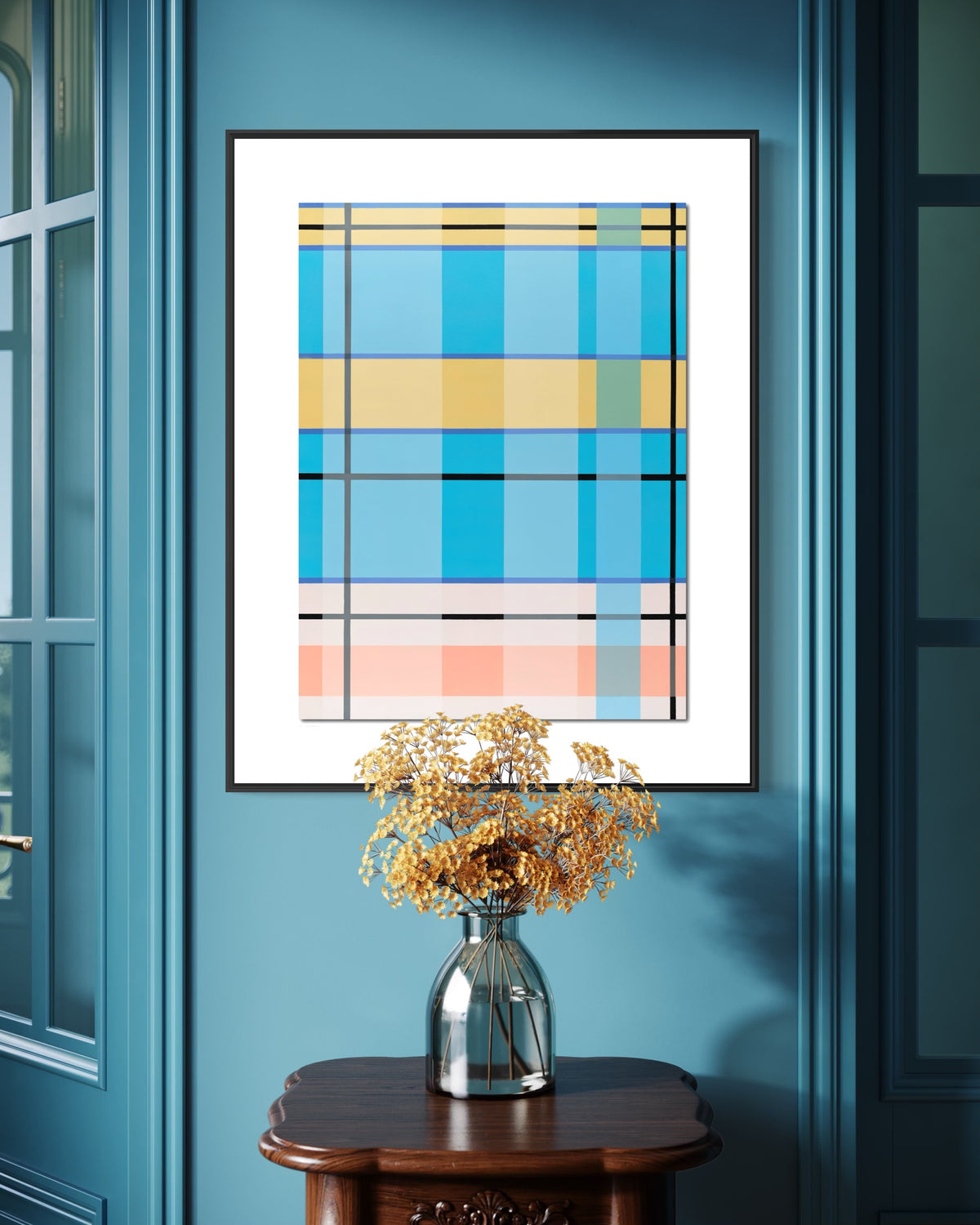 A framed abstract geometric art print with blue, yellow, and pink plaid-like blocks hangs on a teal wall above a dark wood console table with a silver vase of dried flowers.
