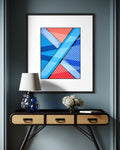 A framed abstract urban art print titled The Search and premised on the search for meaning in life with red, blue, and white shapes hangs on a light gray wall above a red metal chair in a sunlit room.