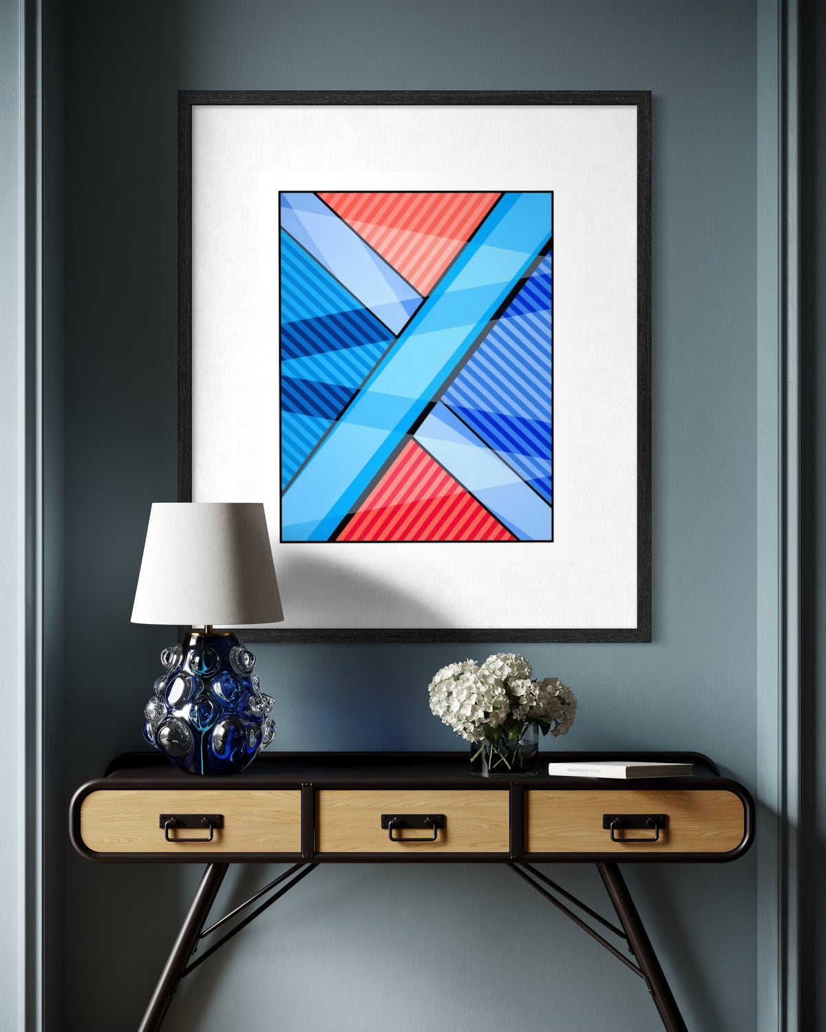 A framed abstract urban art print titled The Search and premised on the search for meaning in life with red, blue, and white shapes hangs on a light gray wall above a red metal chair in a sunlit room.